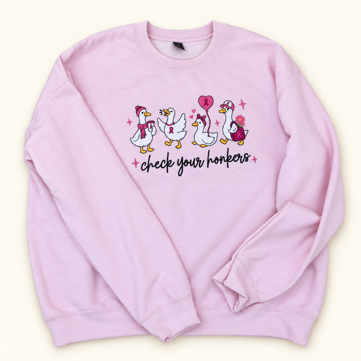 Pale pink Check Your Honkers embroidered crewneck sweatshirt with four geese and pink awareness ribbons on a warm ivory studio background