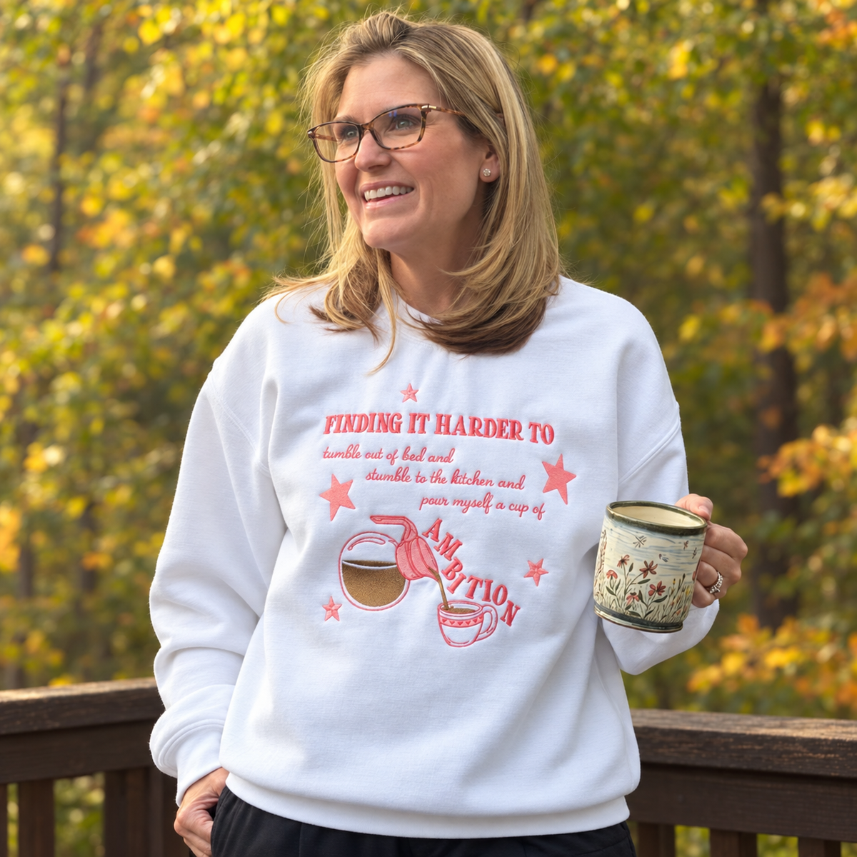 Woman holding a floral mug while wearing a white Cup of Ambition embroidered sweatshirt outdoors among autumn trees