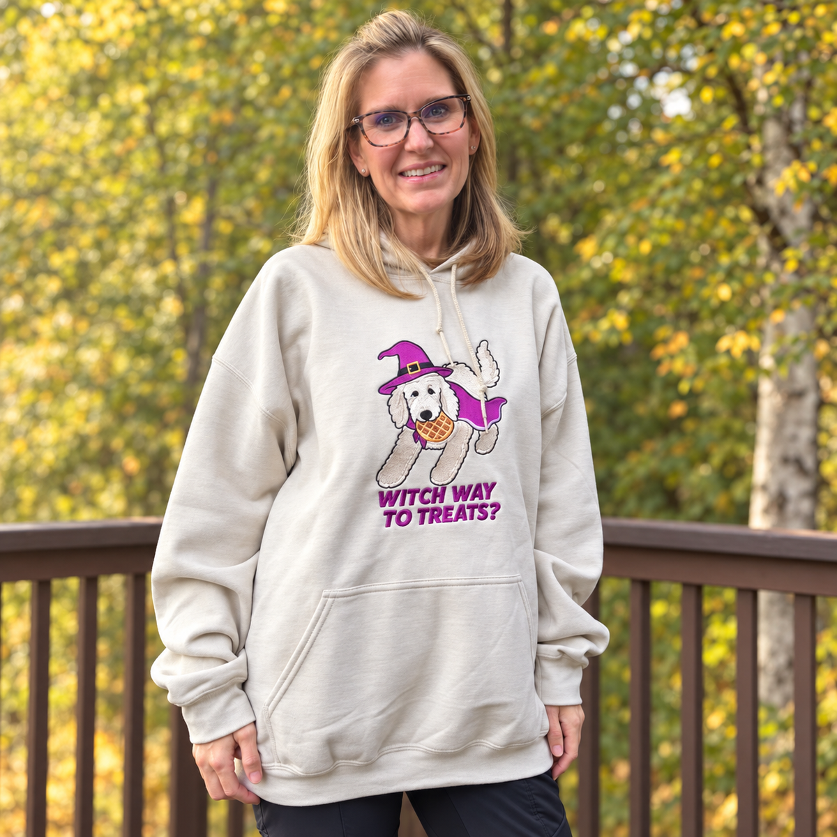 Woman wearing a beige Witch Way to Treats embroidered hoodie outdoors among autumn trees